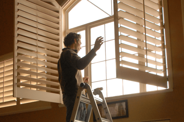 shutter installer standing on ladder while looking out window with open plantation shutters